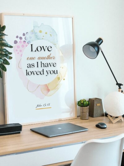 Love One Another Wall Art in a black frame on the wooden desk next to a black lamp and a laptop