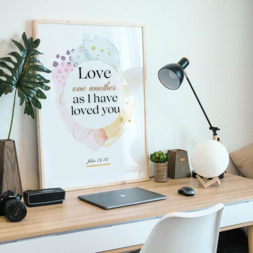 Love One Another Wall Art in a black frame on the wooden desk next to a black lamp and a laptop